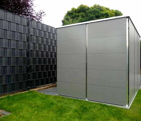 . The image shows a metal garden shed situated on a grassy area. The shed is adjacent to a tall fence with a grid-like design. There are trees in the background.