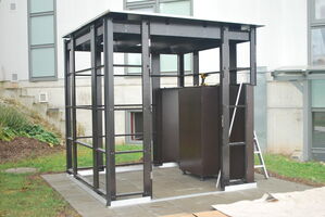 The image shows a metal-framed structure, possibly under construction or for a specific installation. It includes a roof and partial walls, and there appears to be a ladder leaning against it. The structure is positioned on a paved area next to a building, surrounded by grass and landscaping.