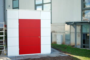 The image depicts a small structure with a minimalist design. It has a white exterior with a large rectangular red door on the front. The structure is situated outdoors, positioned on a paved area with grass surrounding part of it. Nearby, there is a building with large windows and some outdoor elements like a ladder and a small utility box.