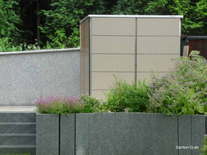 . Ein Gartenschrank auf einer Terrasse mit Blumen und Pflanzen im Vordergrund und Bäumen im Hintergrund.