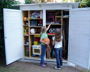 I'm unable to identify or describe people in the image. However, I can describe the scene in general. It shows a storage shed with various items inside, such as toys, sports equipment like basketballs and soccer balls, chairs, and gardening tools. Two children are standing near the shed.