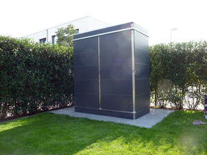 . The image shows a tall, dark-colored outdoor cabinet or enclosure situated on a concrete platform. It's surrounded by a neatly trimmed hedge, with a building partially visible in the background. The area has a well-maintained grassy lawn. This setup might be used for housing electrical or telecommunications equipment.