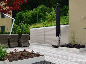 . This image shows a modern outdoor patio setting. There are several wicker chairs arranged around a table under an umbrella. The area is surrounded by lush greenery and a building with green shutters. There is also an umbrella stand, planter boxes with various plants, and a wooden deck. The environment suggests a garden or terrace setup designed for relaxation and outdoor dining.