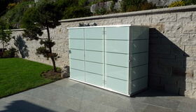 . The image shows a modern storage unit or cabinet with a sleek design, positioned outdoors against a stone wall. The unit has a glass or glass-like appearance with horizontal dividers. It’s set on a stone patio next to a trimmed lawn with a small tree and landscaping in the background.