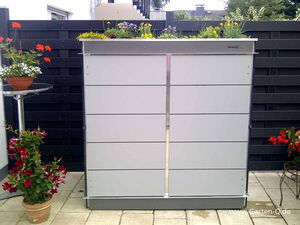 . The image shows an outdoor setting with a modern, sleek storage unit against a dark fence. The unit has a planter on top, filled with various colorful flowers. There are additional potted plants placed on either side of the storage unit, adding to the decorative greenery of the space. A small round table holds another potted plant on the left. Overall, the scene appears to be a landscaped garden or patio area.