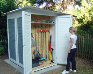 The image shows a person standing in front of an open garden shed. Inside the shed, there are various tools organized on the walls, including rakes, a broom, a shovel, and other gardening equipment. The shed has double doors, and there is a wooden fence and trees in the background.