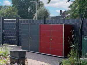 . Ein Garten- oder Geräteschuppen mit roter Verkleidung steht neben einem Zaun, umgeben von Pflanzen.