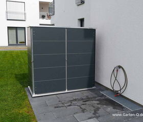 . Eine graue Gartengerätebox steht auf einer Terrasse neben einer Hauswand, mit einem Wasserschlauch an der Wand.