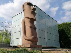 . Eine Moai-Statue steht vor einem modernen Glasgebäude, umgeben von Zaun und Pflanzen.