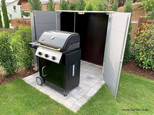 . Ein Grill steht auf einer gepflasterten Fläche zwischen grünen Pflanzen im Garten.