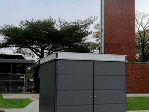 . The image shows a modern, dark gray storage shed or cabinet placed outdoors on a tiled area. There's a tall brick and concrete structure in the background as well as trees. The setting appears to be an open public space or garden.