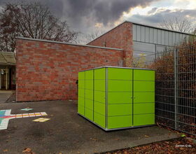 Grüne Gartenbox vor einem Gebäude mit Backsteinfassade und grauem Himmel im Hintergrund.