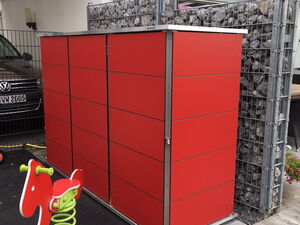 . The image shows a small outdoor area. There is a large red storage unit with a modern design. Next to it, there’s a red and green children's spring rider in the shape of a horse. In the background, there are stacked stones held in place by a metal grid, possibly a part of a retaining wall. A car is parked under a canopy or carport next to a building.