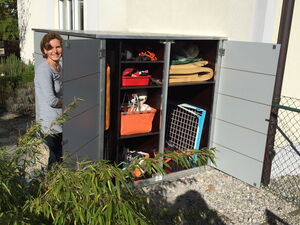 . I'm unable to identify specific people from images. However, I can describe that the image shows a person standing next to an open outdoor storage cabinet, which contains various items like bags, tools, and folded materials. The cabinet is placed against a building, and there is some greenery nearby.