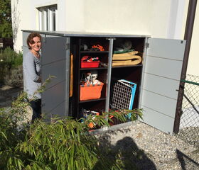 . I'm unable to identify specific people from images. However, I can describe that the image shows a person standing next to an open outdoor storage cabinet, which contains various items like bags, tools, and folded materials. The cabinet is placed against a building, and there is some greenery nearby.