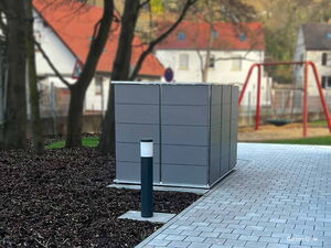 . Ein grauer Gartencontainer steht auf gepflastertem Boden neben einer Lampe, umgeben von Bäumen und Spielplatz.