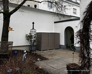 Ein Garten mit Pflanzen, Sitzbank, Vogelhäuschen und einer Box neben einem Gebäude. 