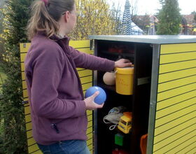 . The image shows a person standing next to an outdoor storage unit with yellow slats. The unit's door is open, revealing various items inside. The person is holding a blue ball and appears to be reaching for something in the unit. Inside, there are items such as a yellow container, a toy truck, and other small objects. The scene is outdoors with trees and other plants visible in the background.