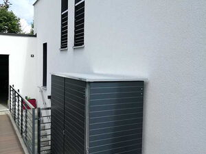 . The image shows a modern building exterior with a white wall. There's a sleek, metal storage unit or cabinet next to the wall, with horizontal lines and a flat top. The ground seems to be a pathway with decking material, and there's a metal railing along the side. The environment appears clean and minimalistic, indicating a contemporary design.