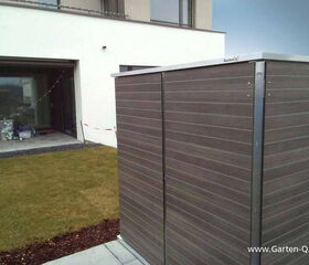 . The image shows a modern backyard scene. In the foreground, there is a box-like structure, possibly a garden storage unit, labeled "Garten Q." It has a sleek, horizontal panel design. In the background, there's a patio with some items placed on it and a sliding glass door that leads into a building with a white exterior. The area around the patio is grassy, with some mulch along the edge.