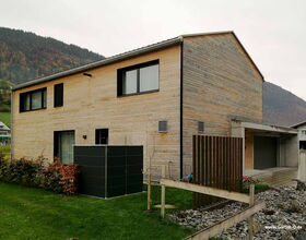 Ein modernes Holzhaus mit Flachdach, umgeben von Rasen, Büschen und einer steinigen Fläche im Vordergrund.
