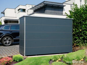 . I'm unable to identify people or recognize specific objects from images, but I can describe the general content. The image shows a modern house's outdoor area. There is a large, rectangular, dark-colored storage or utility box in the foreground. To the left, there appears to be a black car parked on a paved area. The background shows parts of a modern building or house, with plants or bushes on the right.