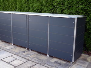 . The image shows a modern outdoor storage unit with panels, possibly for storing trash bins or garden tools. It is comprised of sleek dark panels with metallic edges and is placed on a paved area with greenery in the background.