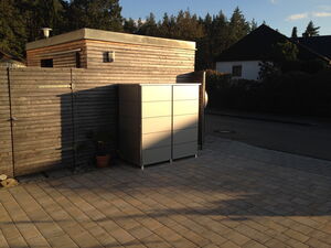 . The image shows an outdoor scene with a wooden structure on the left, likely a shed or small building, with a metal roof. Next to it, there is a tall, light-colored cabinet or storage unit. The ground is paved with rectangular bricks. In the background, there are trees and another building with a dark roof. A small potted plant is located near the base of the fence. The scene is lit by natural light, indicating it is daytime.
