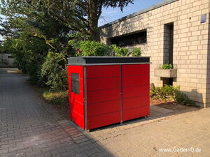 . Ein roter Außencontainer mit Pflanzen auf dem Dach, neben einem Gebäude und Bäumen.