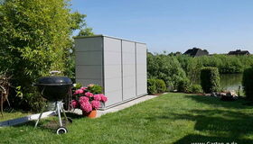 . Garten mit Grill, Pflanzkübel mit Blumen, grauem Schrank, Rasenfläche und Teich im Hintergrund.