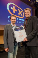 The image shows two people standing on a stage holding a certificate. The background displays a logo for the "Plus X Award." The certificate has text in German, mentioning categories like "Innovation, Design, High Quality."

Here's a description in array format:

- Two people
- Holding a certificate
- Stage background
- Plus X Award logo
- German text on certificate

If you have more specific questions about the image, feel free to ask!