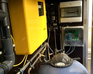 The image shows a section of a utility or equipment room. It features:

1. A large, yellow device mounted on the wall, possibly an electrical component like an inverter or charger.
2. Various pipes and plumbing, some of which are connected to the equipment.
3. An electrical panel or circuit box with protective casing.
4. A large blue pressure tank, possibly for water or air.
5. Wiring and cables running between the devices.
6. A transparent case containing electronic components, possibly for monitoring or control purposes.