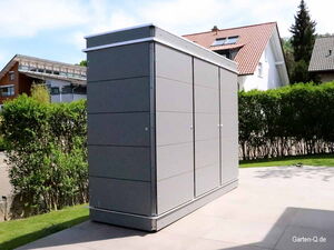 . Ein graues Gartenhaus steht auf einer Terrasse, umgeben von Hecken und Wohnhäusern im Hintergrund.