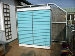 . Ein türkisfarbener Gartenschrank auf einer Terrasse neben einem Gewächshaus und Pflanzentöpfen.