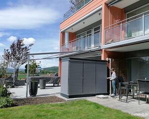 Moderne Terrasse mit Gartenhaus, Möbeln und Pflanzen; Person steht neben dem Gartenhaus.
