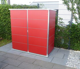 . The image shows a red outdoor storage shed located on a patio. It is positioned against a fence with greenery around and is placed on a paved area with stones and tiles. There is also a small tree nearby.