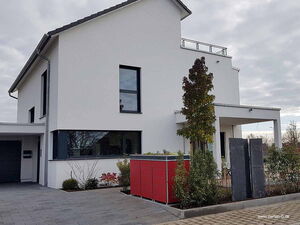 . Modernes weißes Haus mit flachem Dach, großen Fenstern, einem kleinen Garten und roter Box im Vordergrund.