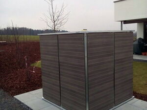 . The image shows a small, modern outdoor storage shed with wooden panels and a metal frame placed on a paved area. It’s situated in a garden or yard area, with a bare tree nearby and a building to the right. The background features a lawn and a field with overcast skies.