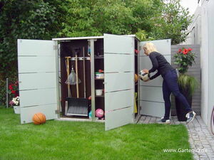. The image shows a person organizing or retrieving items from an outdoor storage cabinet or shed. The cabinet is open and contains various garden tools and sports equipment like rakes, shovels, brooms, and balls. There is also a basketball on the grass. The surroundings include lush greenery and some potted plants with red flowers.