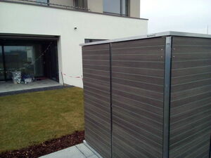 . The image shows an outdoor area with a lawn and a patio. In the foreground, there is a storage shed labeled "Garten Q." The background features a modern building with large windows. The patio has some items stored in clear plastic containers, and there's a decorative light string hanging nearby.