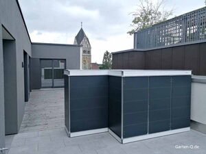 . Moderne Terrasse mit grauen Gebäuden, Holzboden und einem Metallschrank. Im Hintergrund ein Kirchturm.