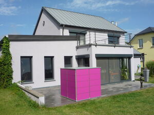 . Ein modernes Haus mit Flachdach und Terrasse, ein markanter pinker Kubus im Vordergrund, dahinter ein weiteres Haus.