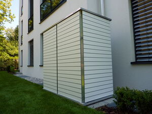 . The image shows a white, modern utility box or shed attached to the exterior wall of a building. It's placed on a patch of green grass and is surrounded by small shrubs. The building has several windows with blinds.