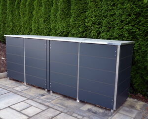 The image shows a large, dark-colored, outdoor storage unit or cabinet. It is placed on a tiled patio area, and there’s a green hedge in the background. The cabinet appears to be made of metal or a similar material, with a design featuring vertical panels and a flat top.