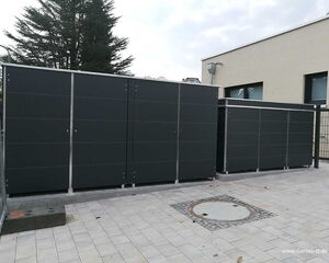 . The image shows several large, dark-colored outdoor storage units or cabinets placed on a paved area next to a building. These units have sleek, modern designs, typically used for storing tools, equipment, or other items. There is a tree in the background, and the area appears to be enclosed by a fence.