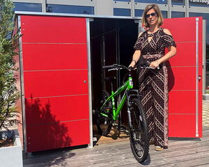 . Frau mit Sonnenbrille holt ein grünes Fahrrad aus einem roten Schuppen.