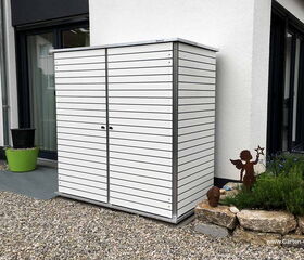 . Ein weißer Gartenschrank steht auf einer Kiesfläche neben einem Haus, mit Pflanzen und einer Engel-Deko daneben.
