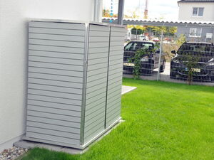 . The image shows what appears to be a heat pump or air conditioning unit enclosure next to a building. It has a sleek, horizontal slat design in a gray color, situated on a green lawn. In the background, there are some parked cars behind a metal fence and neighboring structures.