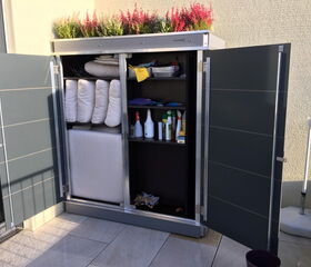 This image shows an outdoor storage cabinet with open doors. Inside, there are various items:

1. Rolled-up blankets or mattresses.
2. Cleaning supplies, such as spray bottles.
3. Other miscellaneous items on the shelves.

On top of the cabinet, there are potted plants with pink and red flowers.