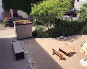 . The image shows a small outdoor area with several elements:

1. A paved area with a bench.
2. A sandy play area with a small picnic table and benches.
3. A rectangular planter with plants on top.
4. Various greenery including bushes and trees.
5. A building with ivy covering the wall.
6. Some fencing and a parked car in the background.
7. An umbrella partially visible at the bottom.